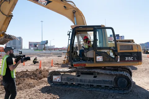 A camera operator filming a host at the controls of an excavator